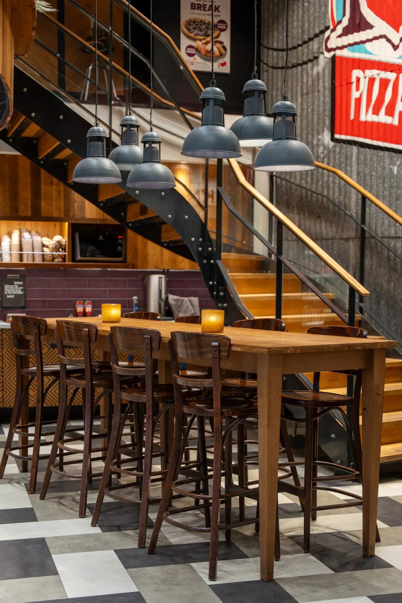 Interiör i en restaurang med lång träbord, höga barstolar, hängande grå industrilampor och en trappa i bakgrunden. Väggen har en stor Pizza-skylt.