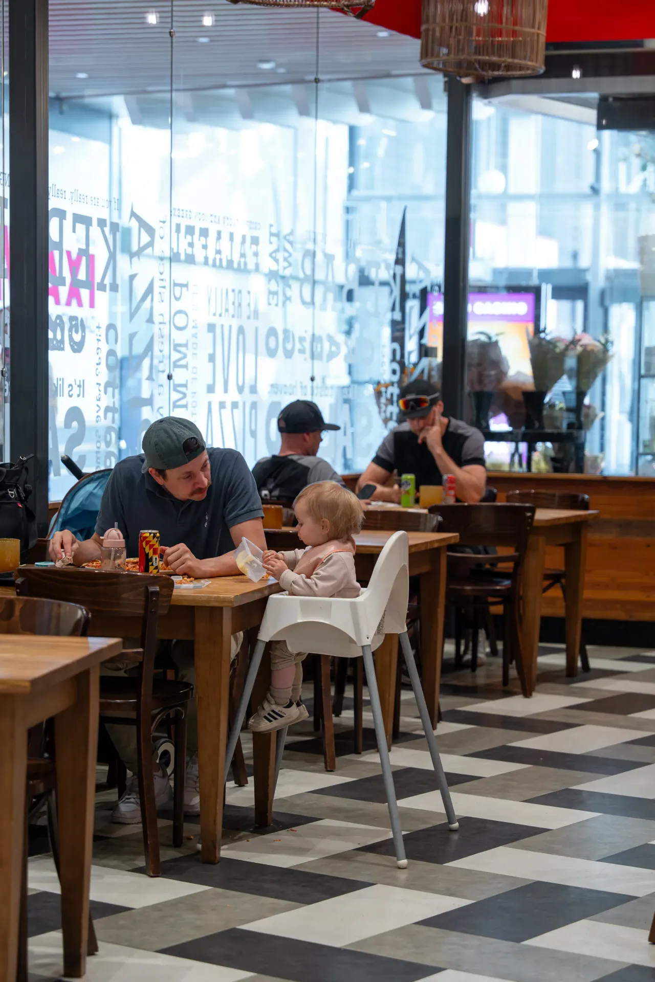 En man äter vid ett bord i en restaurang medan ett litet barn sitter i barnstol mittemot och håller en matburk. I bakgrunden syns fler gäster och stora fönster.
