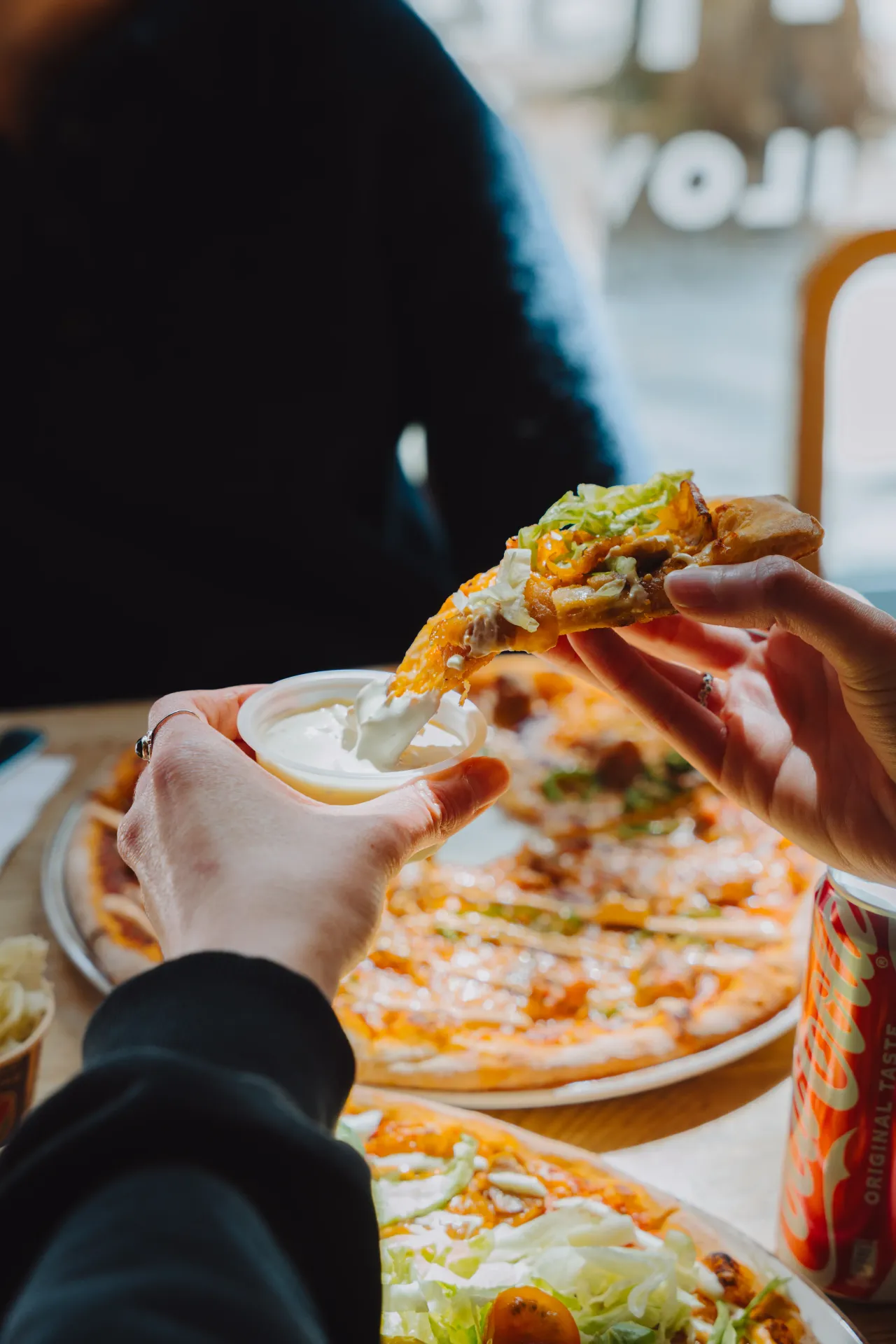 Närbild av händer som doppar en bit pizza med sallad i en liten bägare med sås vid ett bord med flera pizzor och en läskburk.